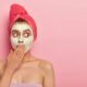 shocked young woman applies clay nourishing facial mask covers mouth with palm hydrates calms skin wears rosy towel wrapped head stands against pink wall rejuvenation concept 80x80